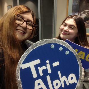 Jennifer and Leah holding a cardboard circle with the words "Tri-Alpha" painted on it
