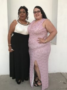Two individuals in dresses hugging and smiling at camera