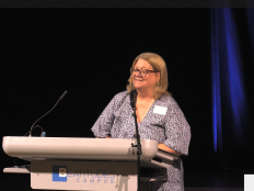 A photograph of Tena Alonzo who has medium length blonde hair and wearing glasses, giving the land acknowledgement at the LGBTQ2S+ Aging Equity Forum