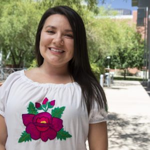 Amanda Hunter who has long brown hair, is wearing a white shirt with a pink flower on it, against a city street backdrop