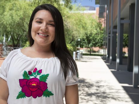 Amanda Hunter who has long brown hair, is wearing a white shirt with a pink flower on it, against a city street backdrop