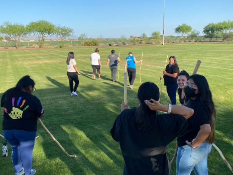Dr. Amanda Hunter learns how to play toka with Salt River Teens. She stands beside 8 others who are listening on a field carrying sticks similar to field hockey sticks.