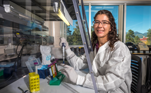 Emily Cope smiles in her lab. She is wearing a white lab coat and is wearing glasses and latex gloves and holding a dropper and a test tube. 