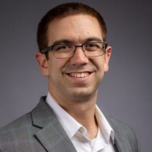 A photo of Eric Cerino, who is wearing a grey jacket and against a grey background. He has short brown hair and is wearing glasses. 