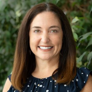 A headshot of Monica Lininger who has medium, straight brown hair, is smiling, and is against a green treed background.