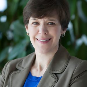 A portrait of Julie Baldwin smiling. She has short brown hair and is wearing a brown jacket and a blue sweater.