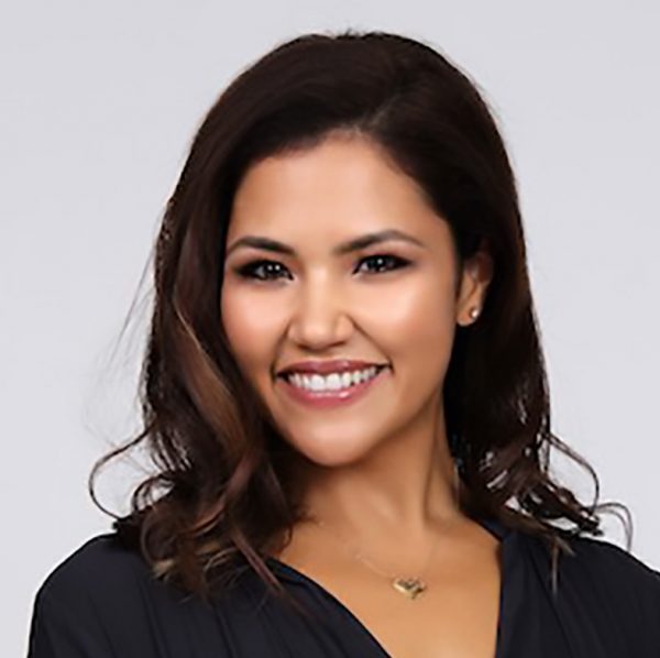 Misty Pacheco poses for a portrait. She has long, dark hair that is curled and is wearing a black v-neck shirt. She is smiling.