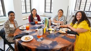 Five women sit around a round table covered with a tie dye tablecloth. On the table are white dishes and clear glasses.