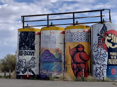 Barrels on the Navajo Nation that are painted with graffiti art, which includes birds and an arm pointed vertically with a hand in a fist that says "Power to the Patients." 