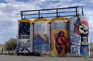 Barrels on the Navajo Nation that are painted with graffiti art, which includes birds and an arm pointed vertically with a hand in a fist that says "Power to the Patients." 
